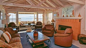A spacious living room with a high ceiling, wooden beams, a fireplace, and a large window overlooking the ocean.