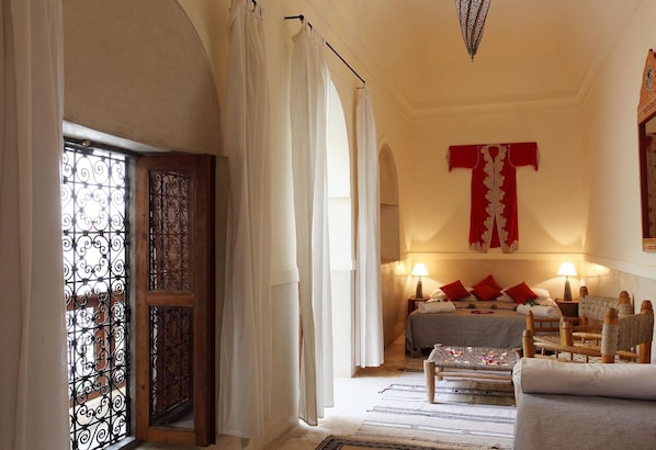 Una habitación con cama, sofá, mesa y lámpara. Cuenta con una ventana con cortinas y una celosía metálica decorativa.
