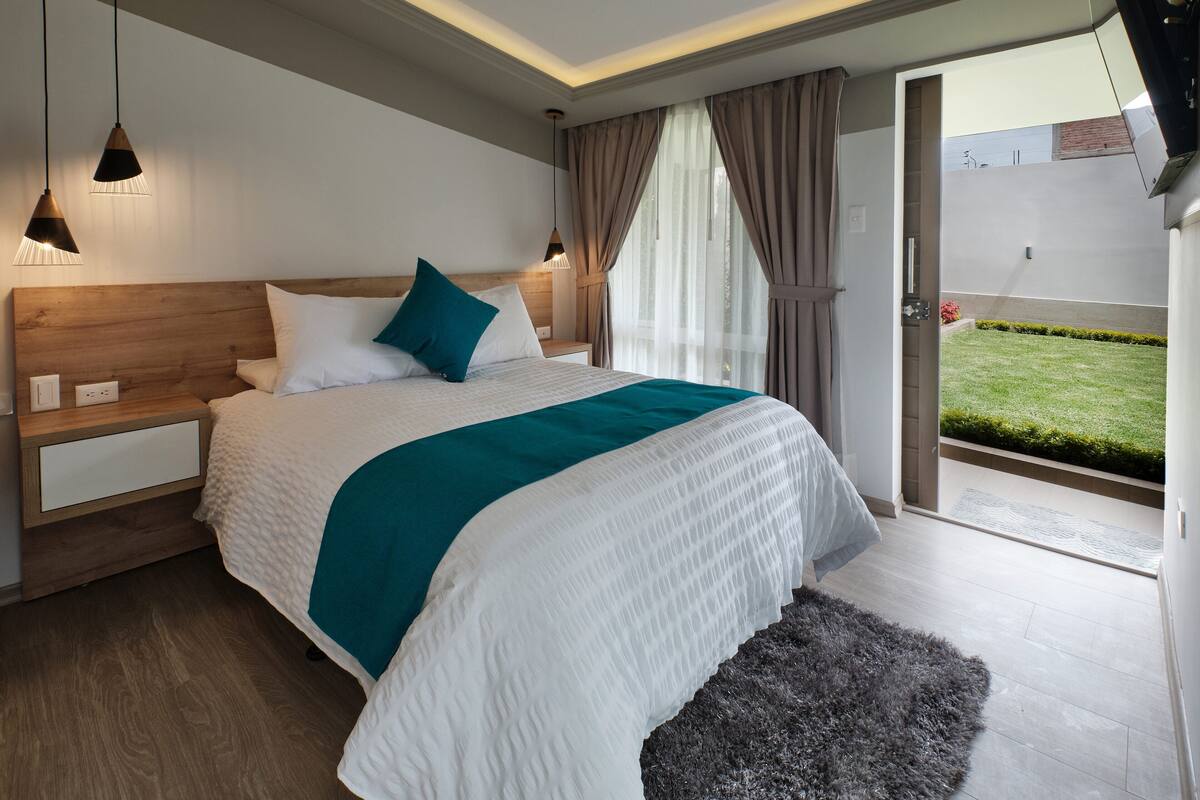 A modern bedroom with a wooden headboard, a large bed with white and teal bedding, a dark rug, and a view of a green lawn through a glass door.