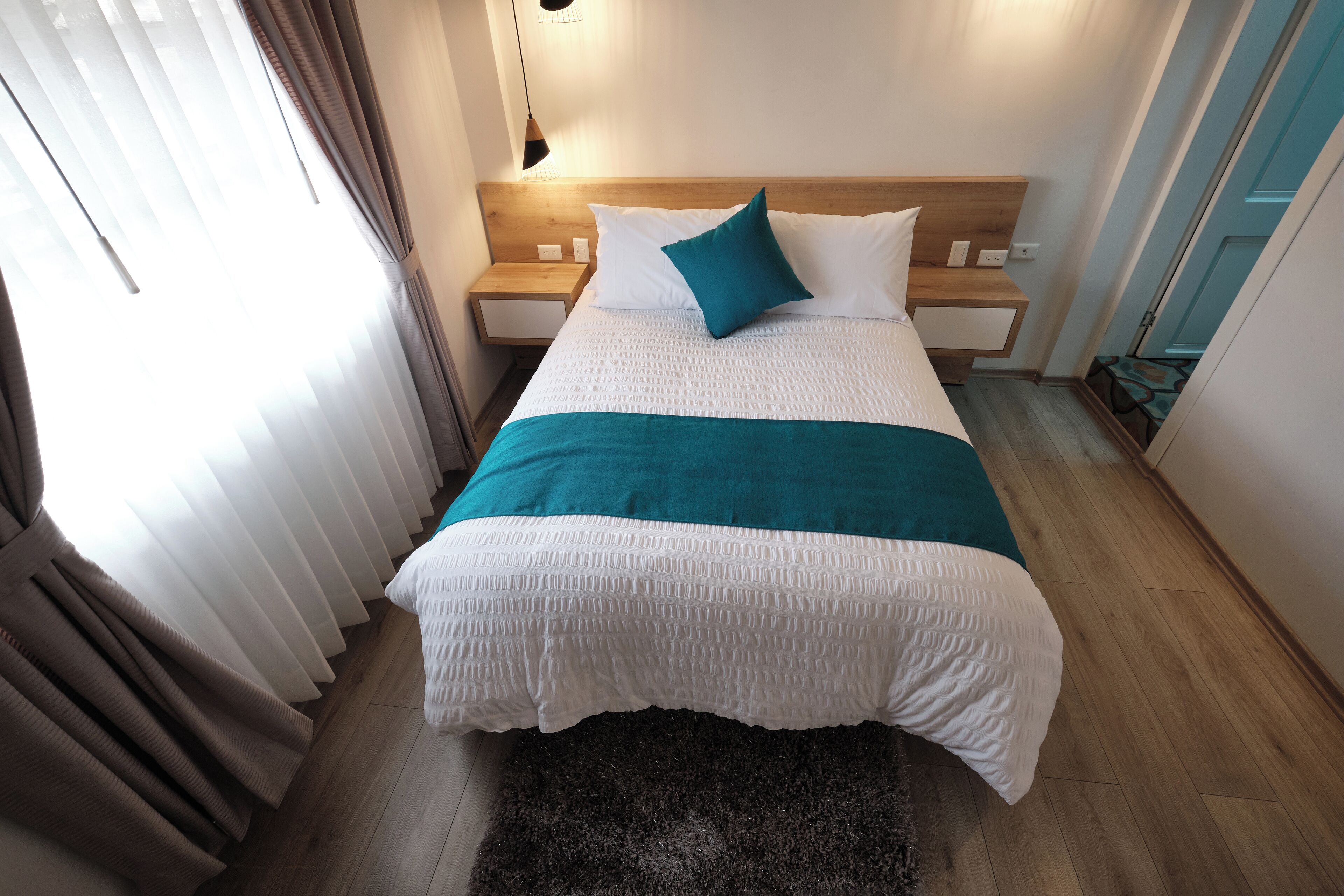 Una cama bien hecha con ropa de cama blanca y aguamarina, un cabecero de madera y una mesita de noche pequeña con una lámpara.