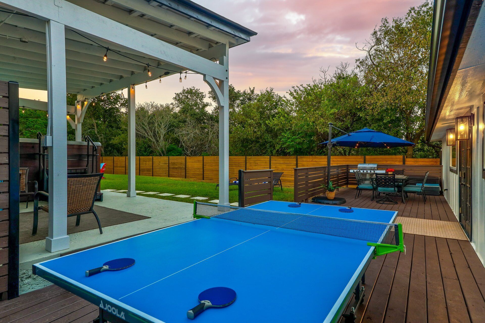 Une table de ping-pong extérieure avec deux raquettes, un barbecue et un espace de détente couvert avec des chaises et des tables.