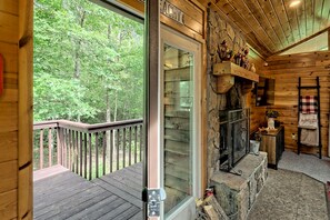 A wooden cabin interior with a glass door leading to a deck, a stone fireplace, and a shelf with decorative items.