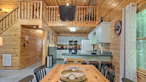 A wooden cabin interior with a dining area, kitchen, and a staircase leading to a loft.