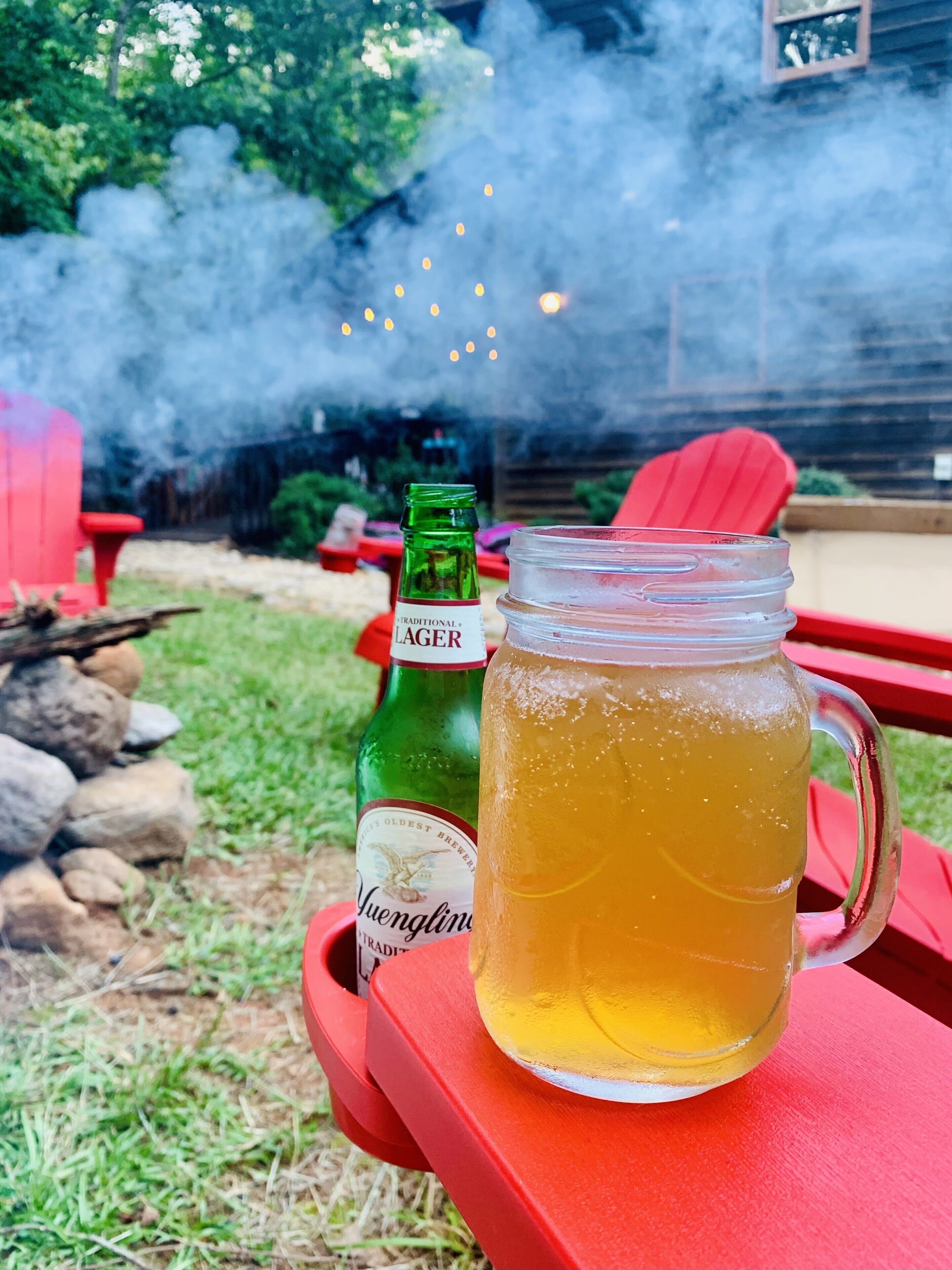 Une tasse en verre contenant une boisson, posée sur une table rouge, avec une bouteille verte et une chaise rouge, le tout dans un décor de jardin.