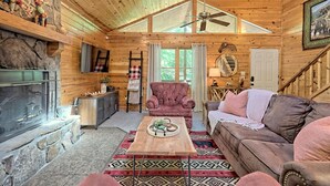 A cozy log cabin interior with a fireplace, plush seating, and a ceiling fan.