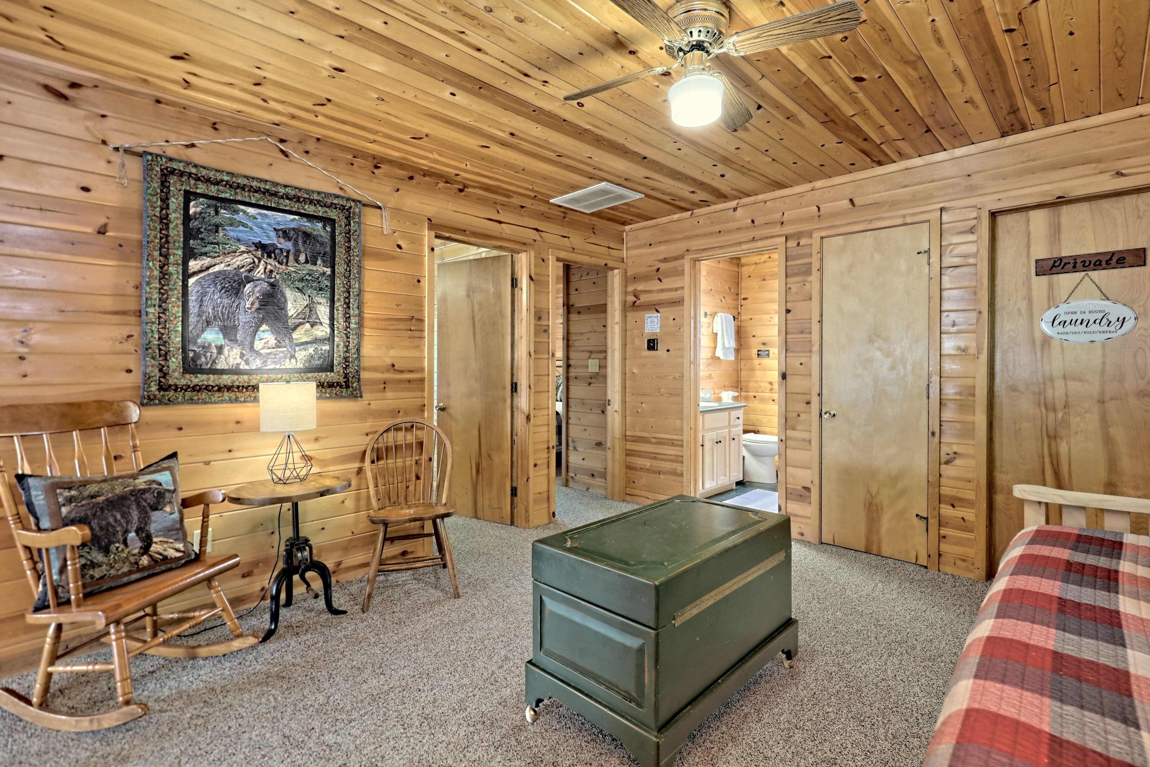 A wooden cabin interior with a rocking chair, a table, a green chest of drawers, a framed wildlife picture, and a sign indicating a private laundry room.
