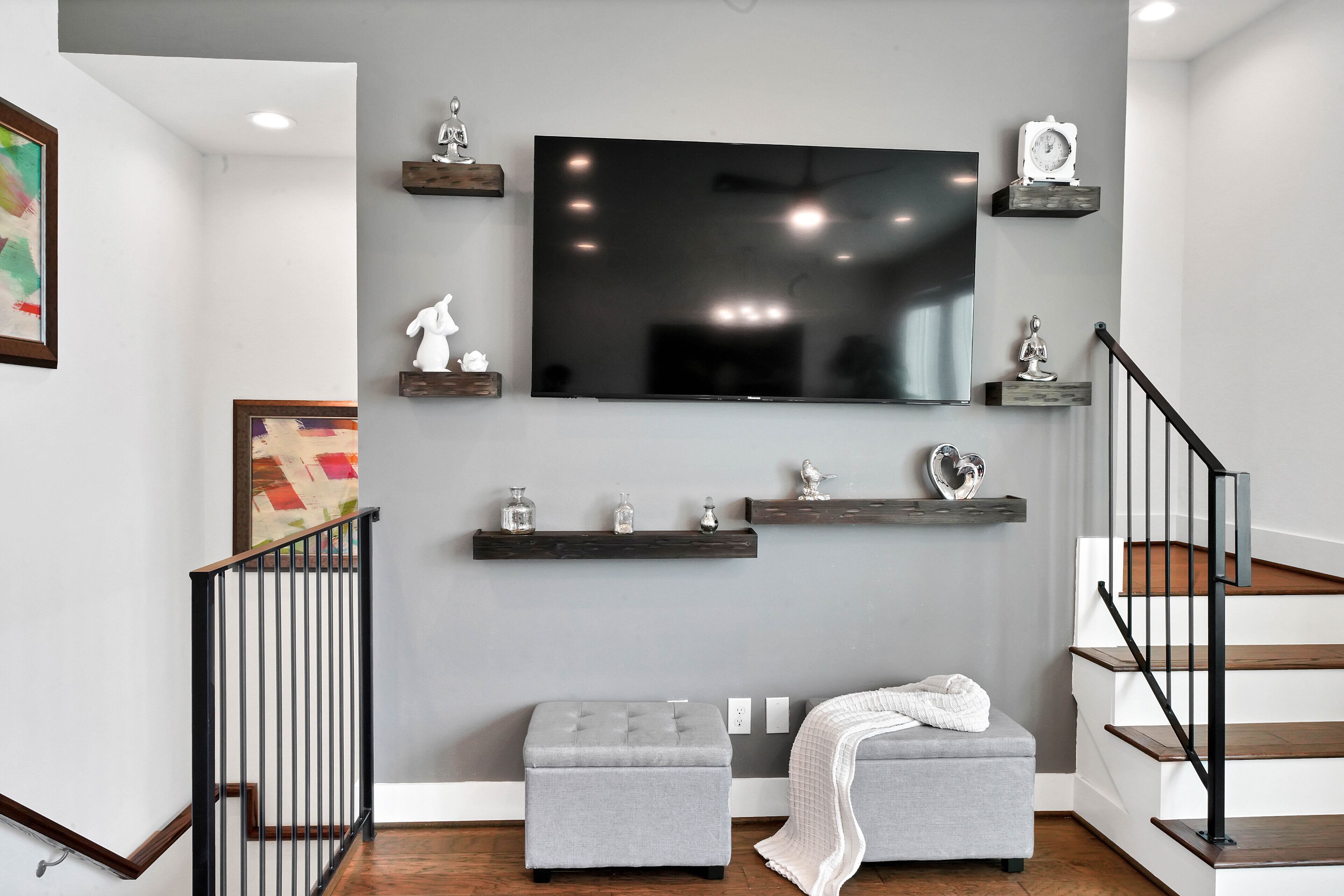 Una sala de estar moderna con un sofá gris, un reposapiés gris, un televisor de pantalla plana montado en la pared y una escalera con barandillas negras.