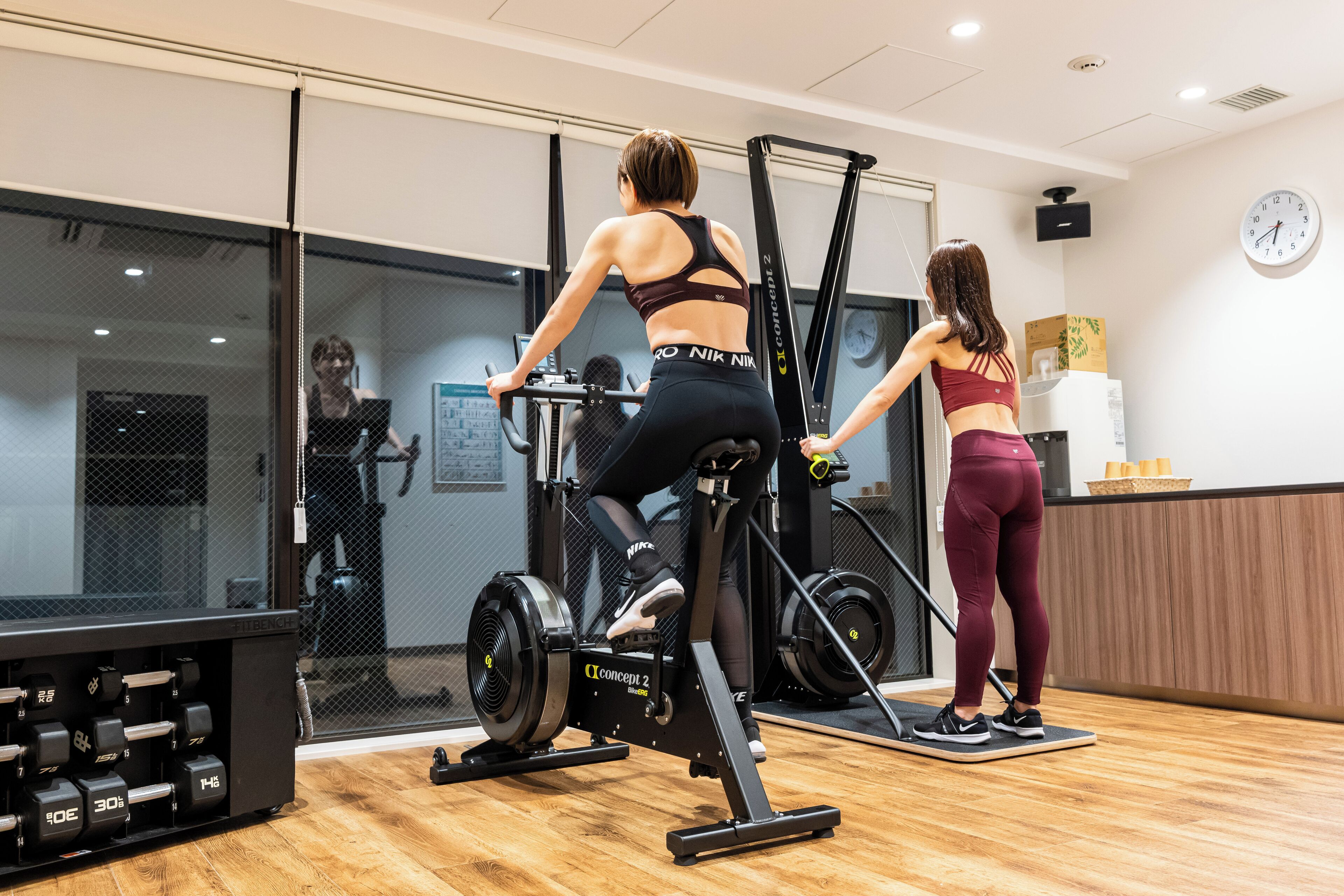 Academia com equipamentos de exercício, um relógio na parede e piso de madeira.