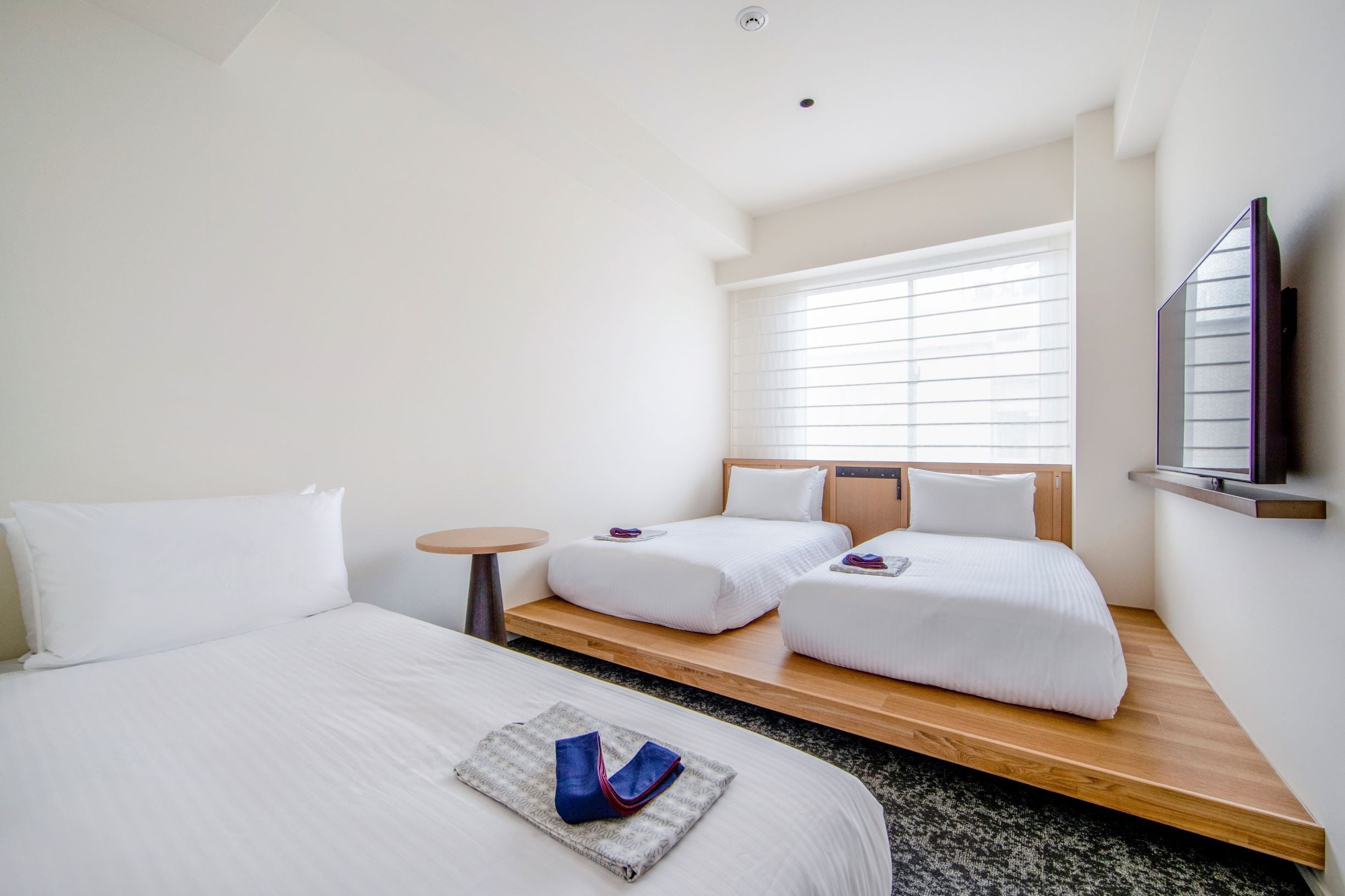 A hotel room with two beds, a TV, a small table, and a window with blinds.