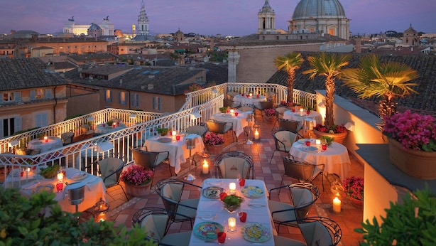 A rooftop terrace with tables set for dining, surrounded by potted plants and candles, overlooking a cityscape with historic buildings and domed structures. - Bio Hotel Raphael - Relais Chateaux (Rome)