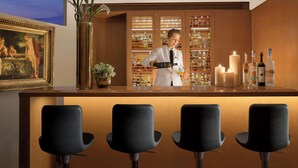 A bar area with a bartender, bar stools, a framed painting, a vase with flowers, and shelves with bottles. - Bio Hotel Raphael - Relais Chateaux (Rome)