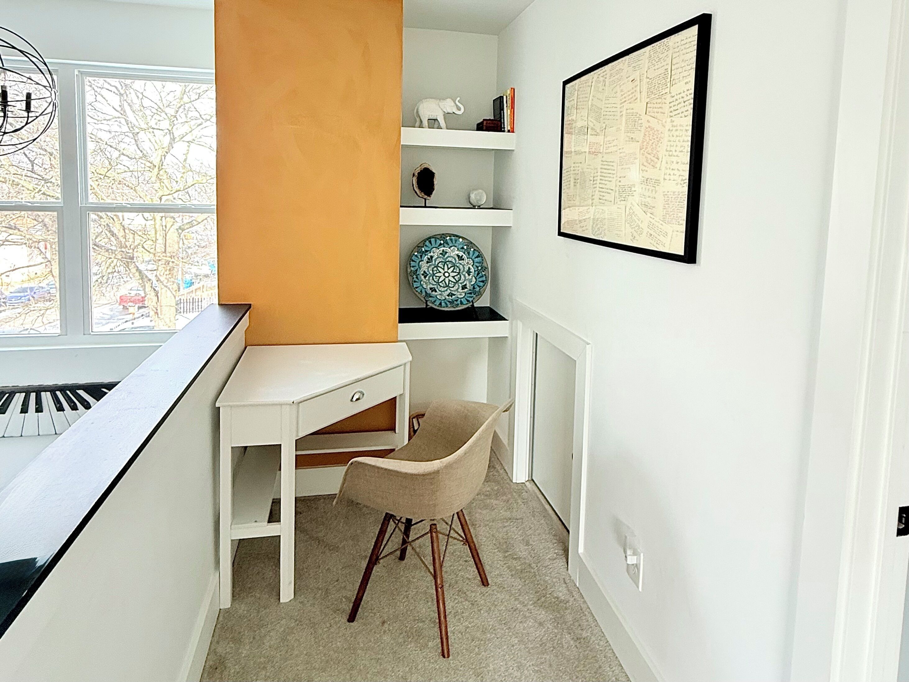 A room with a piano, a desk, a chair, a shelf with decorative items, and a framed document on the wall.
