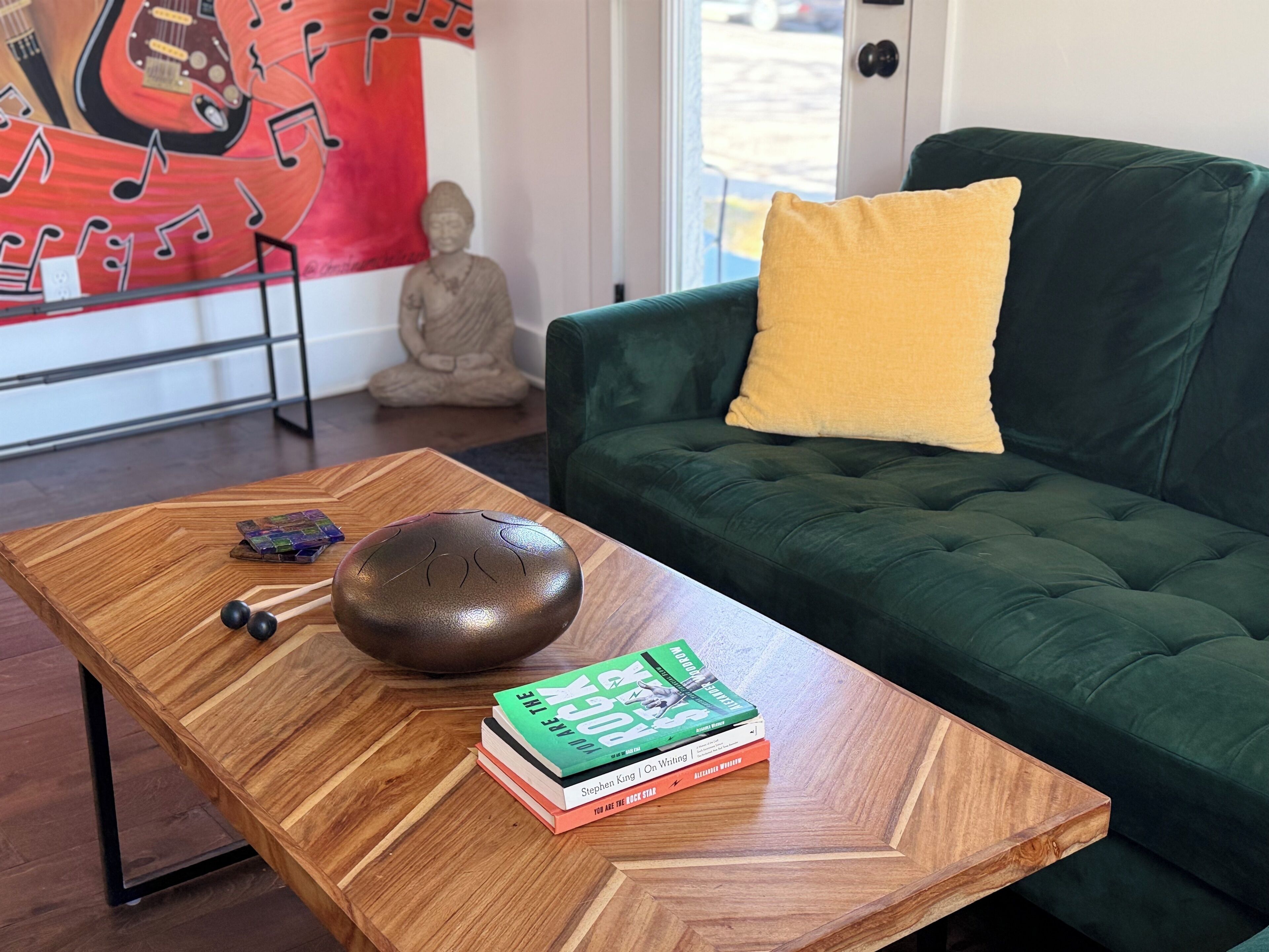 A wooden coffee table with books and a speaker, a green sofa with a yellow pillow, and a Buddha statue in the background.