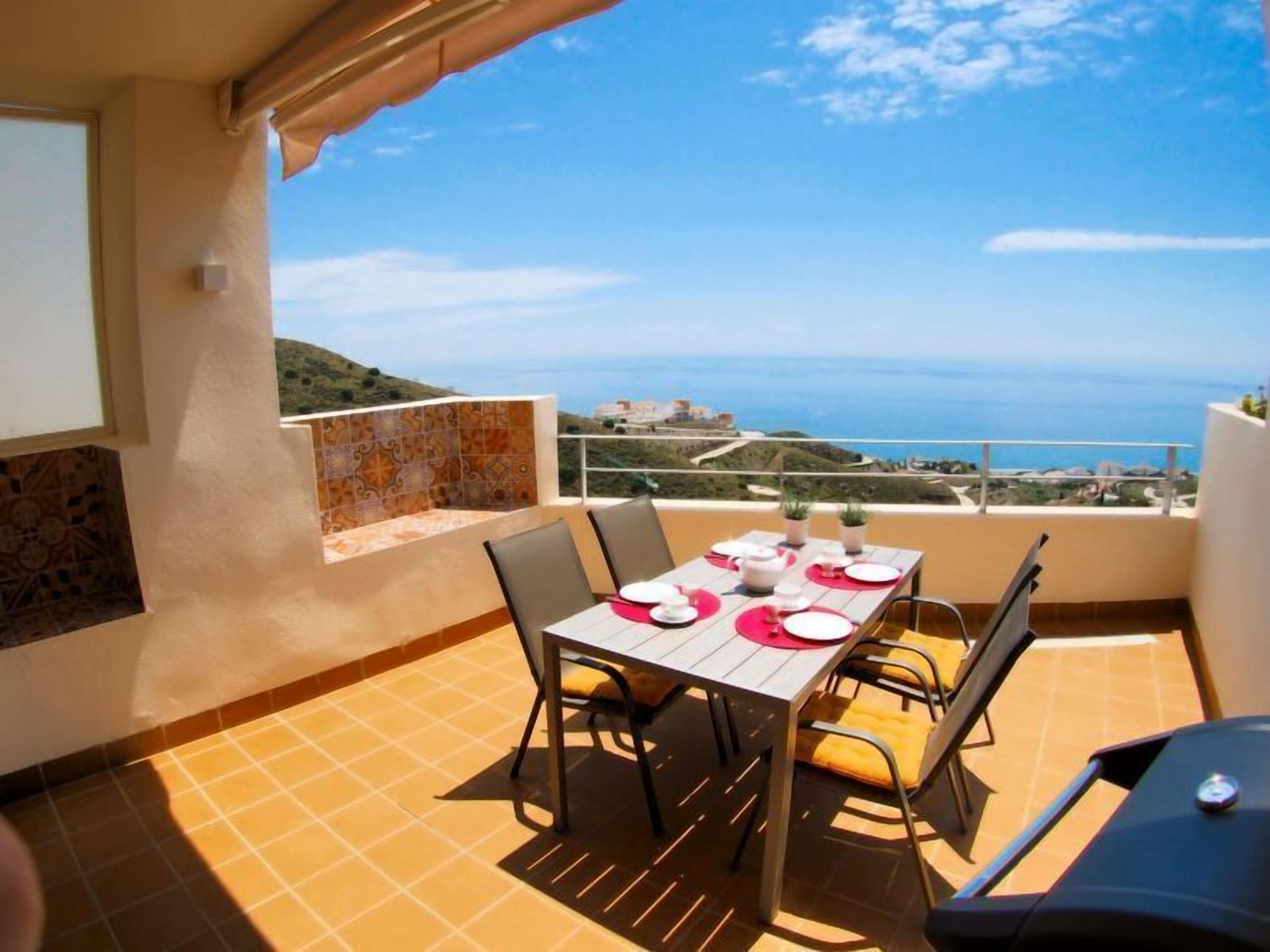 A balcony with a table set for two, overlooking a coastal view.