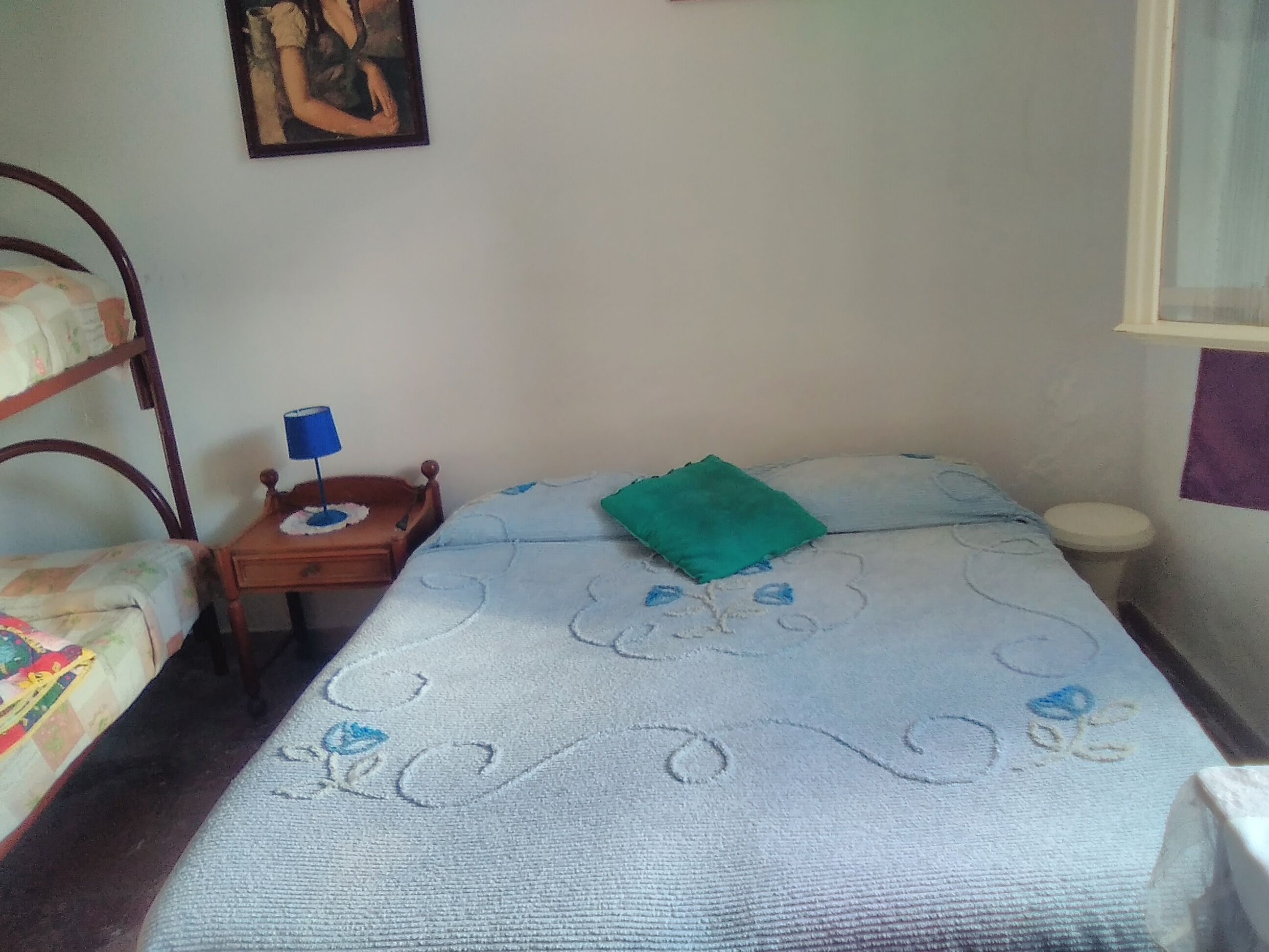 Una habitación pequeña y sencilla con una cama, una mesita de noche, una silla y un estante en la pared.