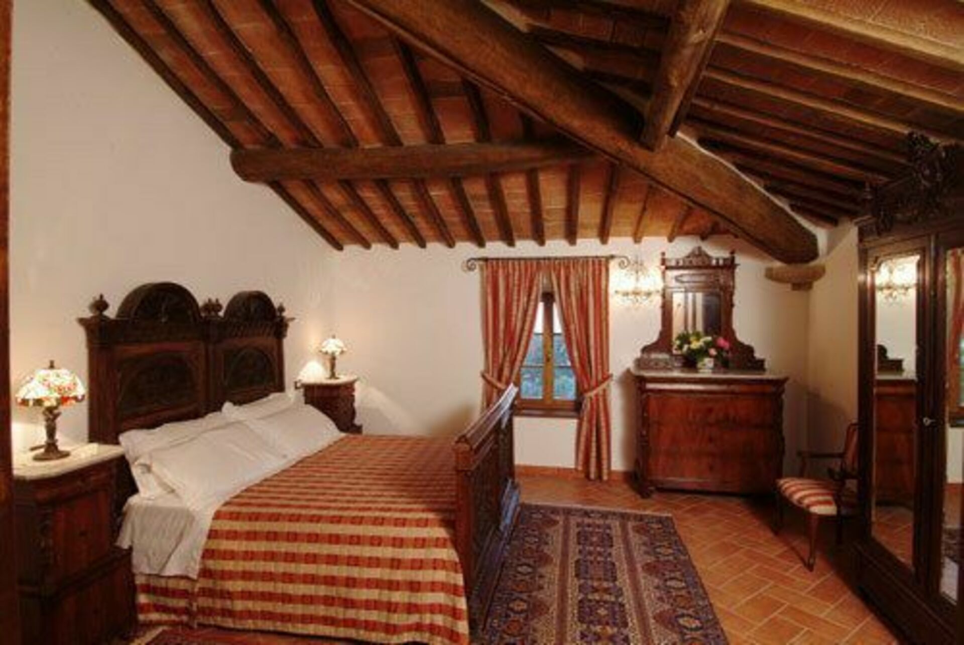 Une chambre avec un plafond en bois, un lit recouvert d’une couverture à carreaux, une table de chevet avec une lampe, une commode avec un miroir et une fenêtre avec des rideaux.