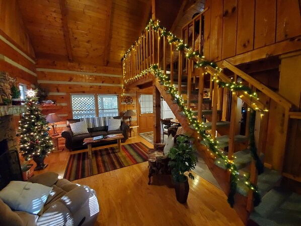 A cozy cabin interior with a wooden staircase decorated with lights, a fireplace, a sofa, and a Christmas tree.