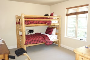 Ein Stockbett mit einer rot-schwarz karierten Decke, einem Kissen mit Bärenmotiv und einem Fenster mit Jalousien.