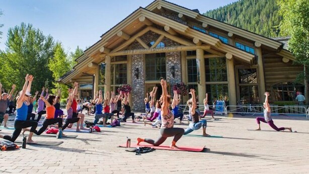 Un grupo de personas practicando yoga al aire libre frente a una cabaña con fachada de piedra y vigas de madera.
