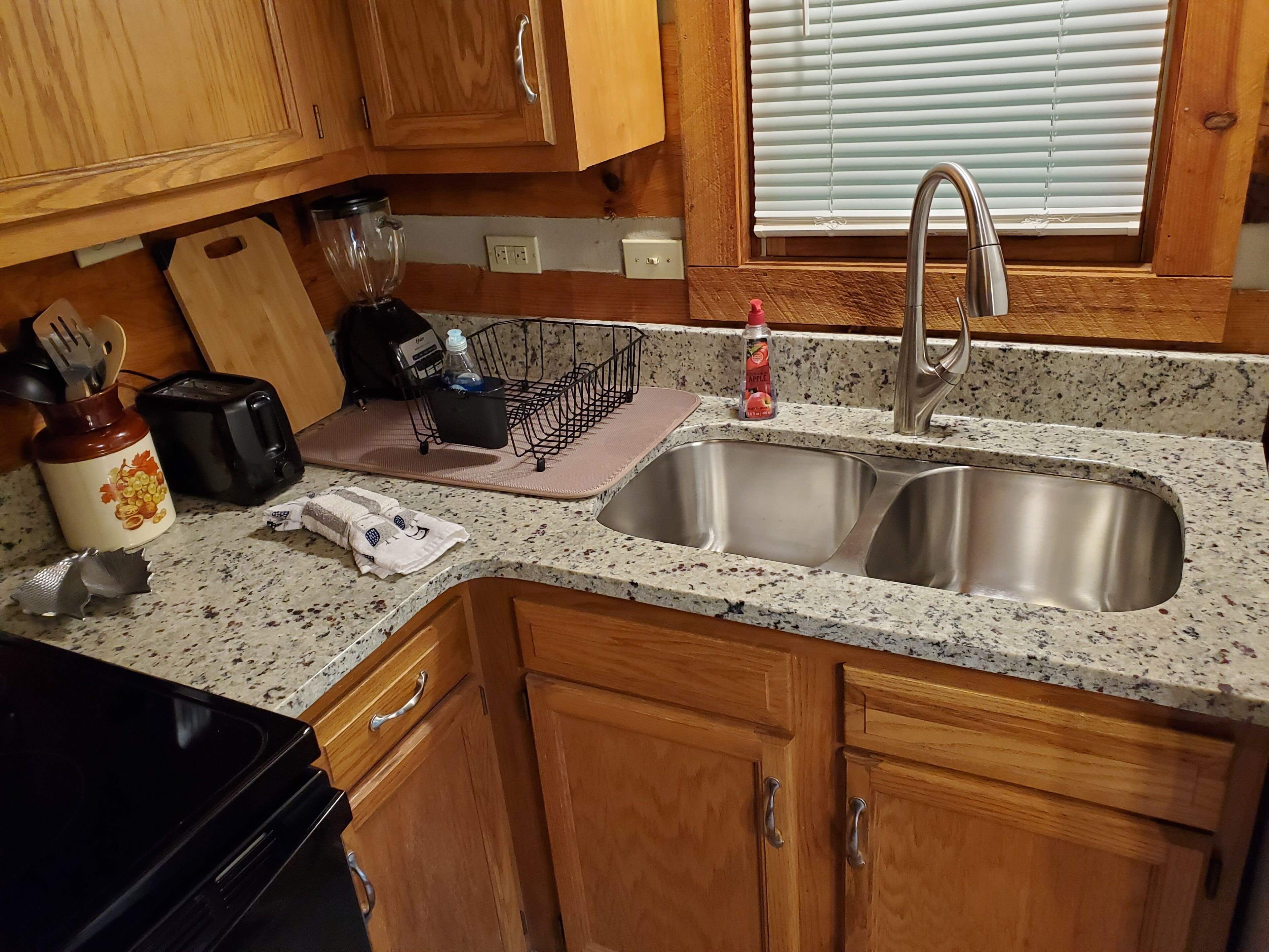 A kitchen with wooden cabinets, a granite countertop, a stainless steel sink, a toaster, a blender, and a cutting board.