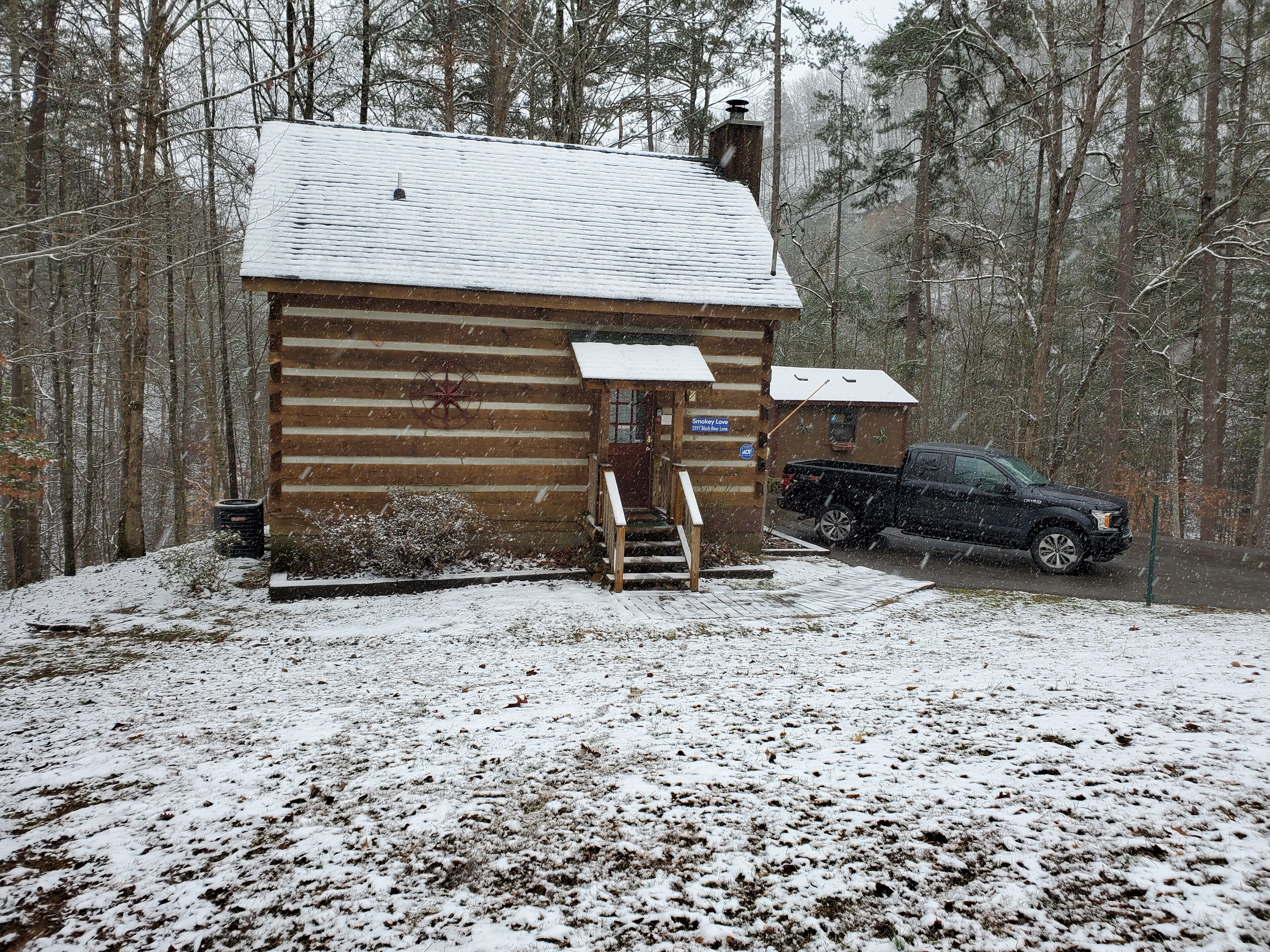 A log cabin with a snow-covered roof, a wooden staircase leading to a door, and a truck parked in front.