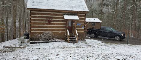 A log cabin with a snow-covered roof, a wooden staircase leading to a door, and a truck parked in front.
