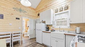 A compact kitchen with white cabinets, a refrigerator, a sink, and a stove. There is a dining table with chairs and a window with curtains. - Riverfront Cottage / Fish Camp Getaway (Newland)