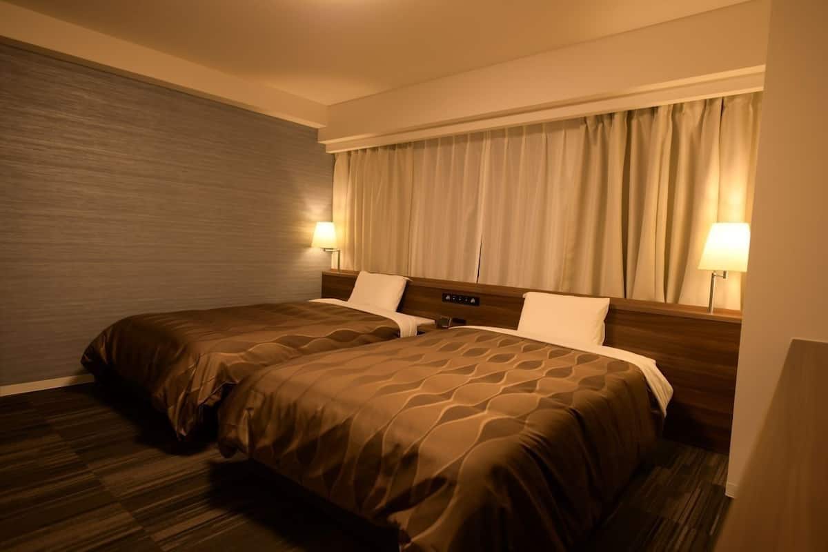 Une chambre d’hôtel avec deux lits, une tête de lit en bois, une grande fenêtre avec des rideaux et deux lampes de chevet.