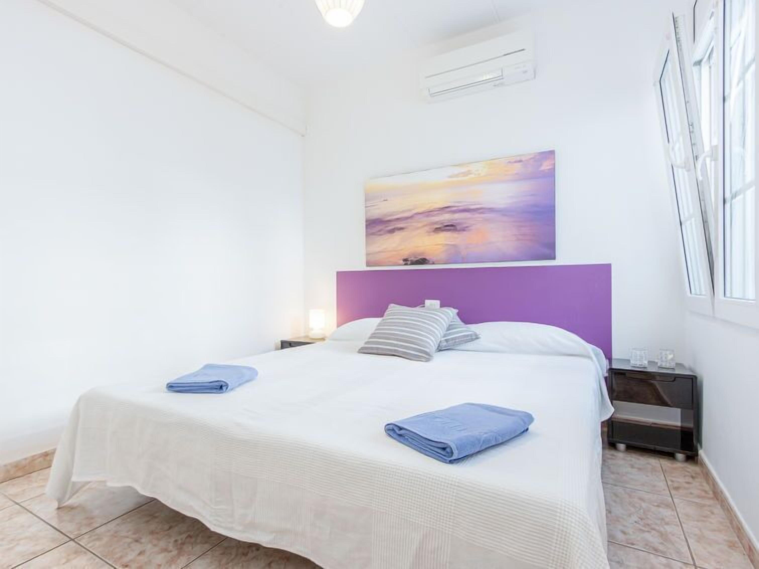 Een moderne slaapkamer met een wit bed, een paars hoofdeinde en een zonsondergangschilderij aan de muur.