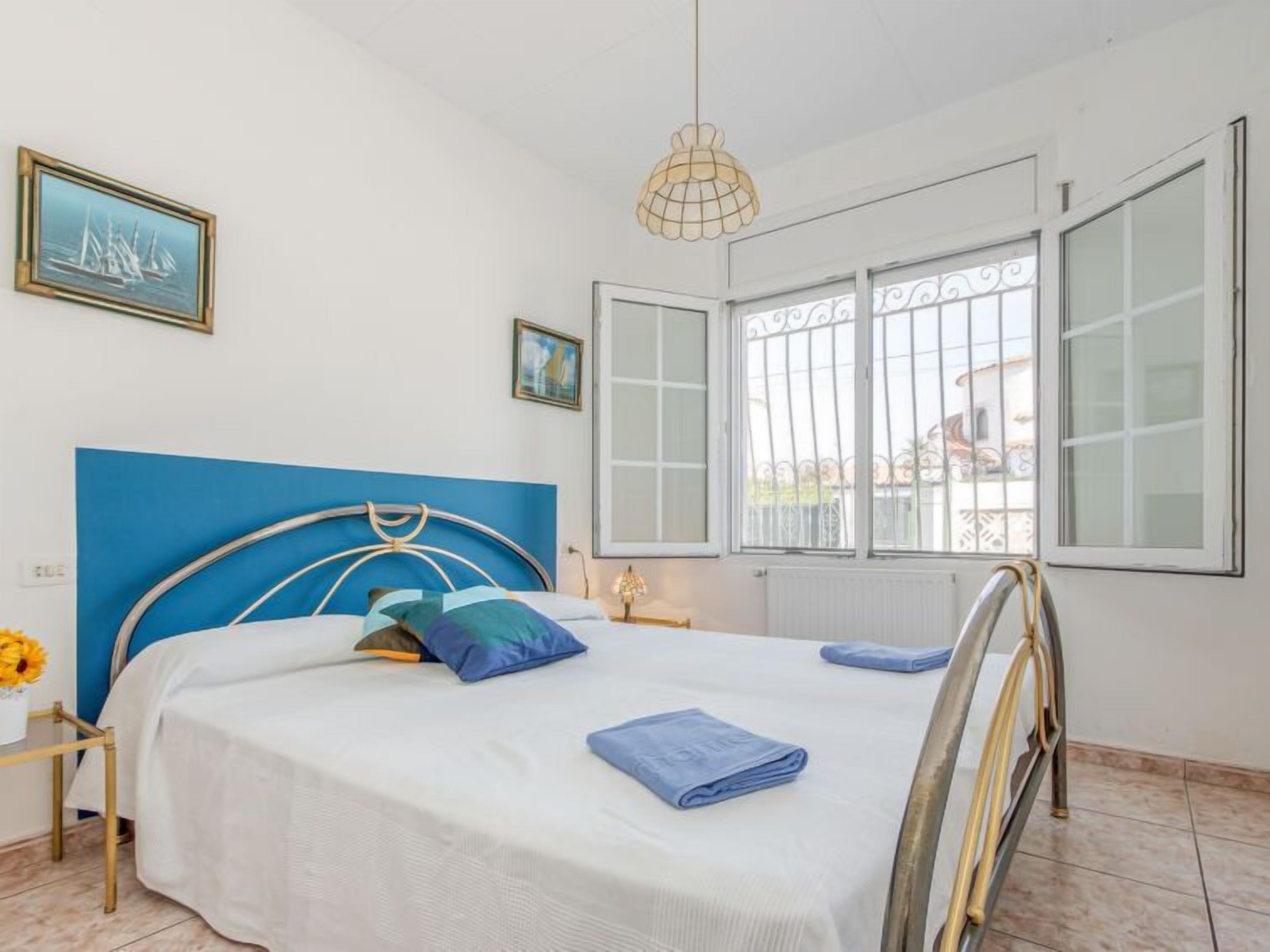 Une chambre à coucher avec une tête de lit bleue, un lit avec une literie blanche, une table de chevet avec une lampe et une fenêtre avec des barreaux en métal.