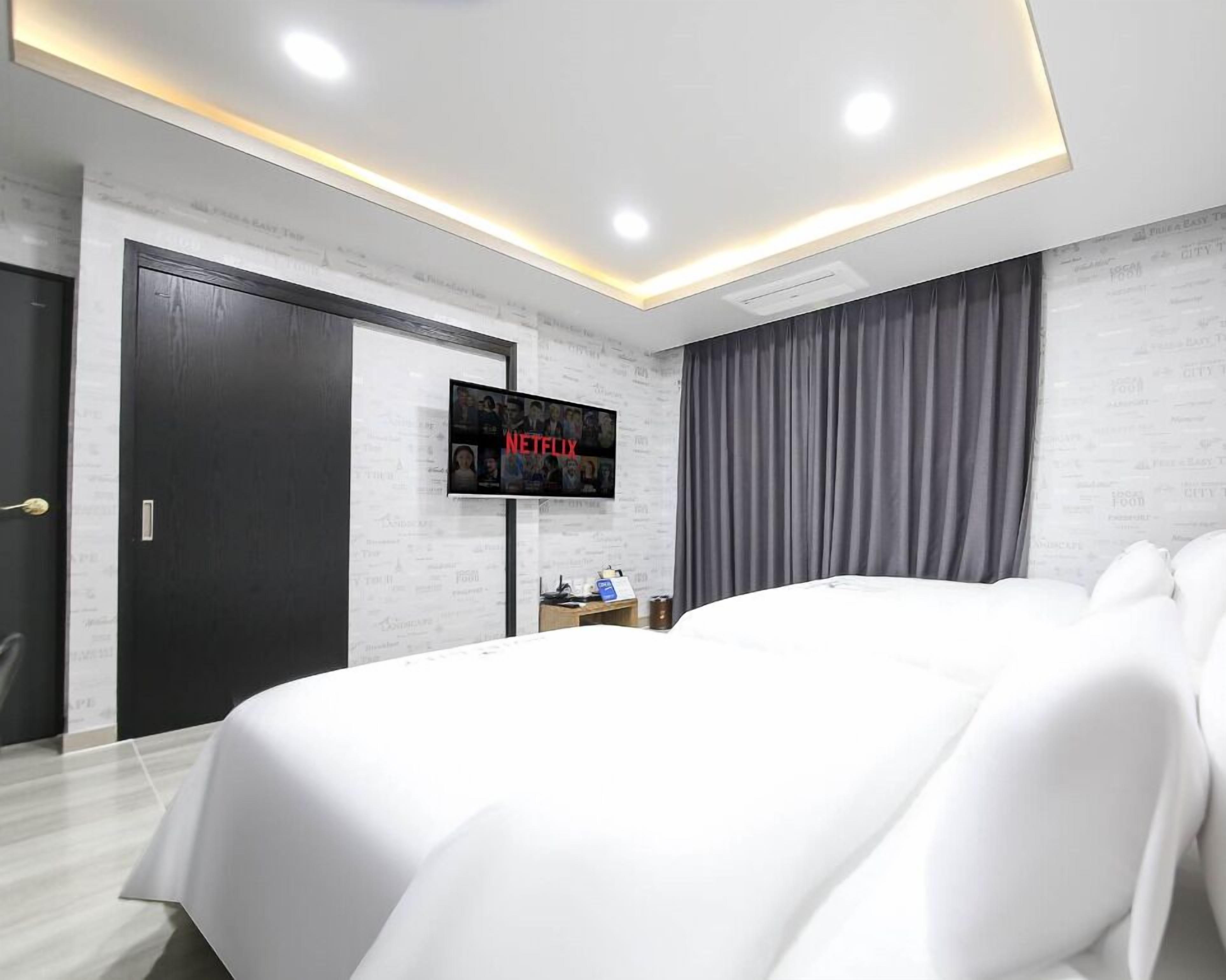 A modern hotel room with a large bed, a flat-screen TV mounted on the wall, a desk with a chair, and a wardrobe.