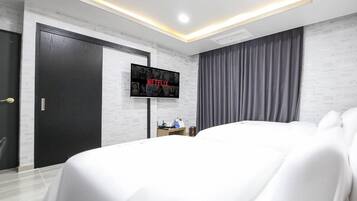 A modern hotel room with a large bed, a flat-screen TV mounted on the wall, a desk with a chair, and a wardrobe.