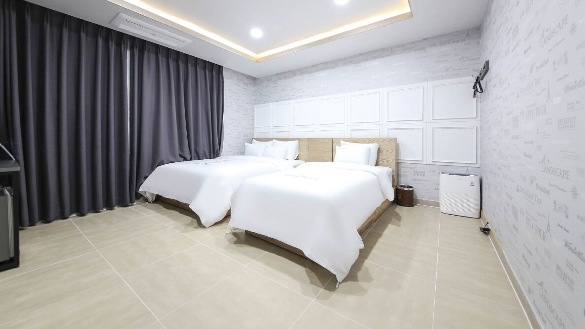 Una habitación de hotel moderna con dos camas, un ventanal con cortinas, un aire acondicionado integrado y un televisor montado en la pared.