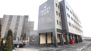 Um hotel moderno com um destaque para a placa "HOTEL CITY", com fachadas de vidro e concreto e um estacionamento na frente.