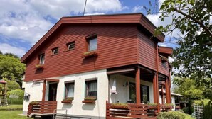A two-story building with a red exterior and white walls, featuring a covered porch with wooden railings and potted plants.