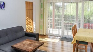 A living room with a sofa, a coffee table, and a dining table with chairs. There is a large window with curtains and a view of greenery outside.
