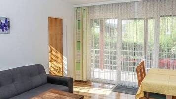 Ein Wohnzimmer mit einer Couch, einem Couchtisch und einem Esstisch mit Stühlen. Es gibt ein großes Fenster mit Vorhängen und einen Blick ins Grüne.
