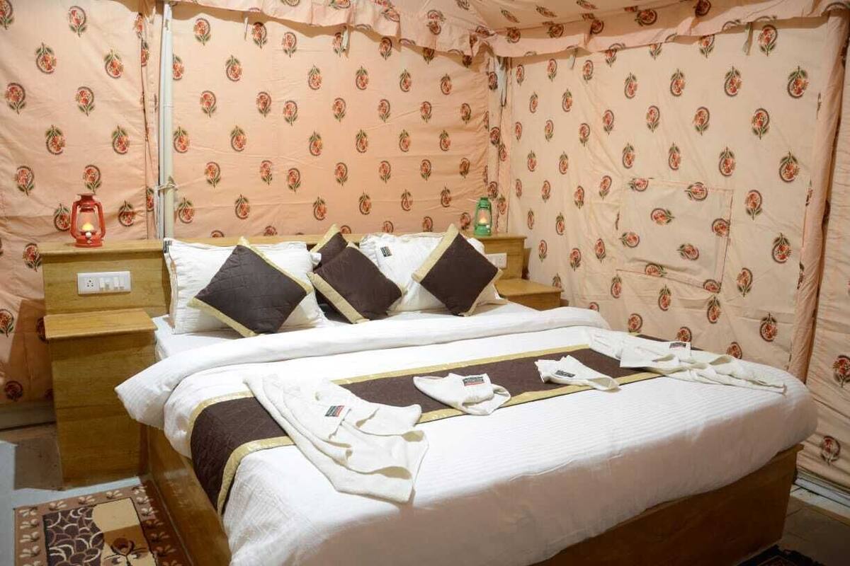Una cama bien hecha con una colcha blanca y marrón, dos almohadas y una mesita de noche con una lámpara y una botella.