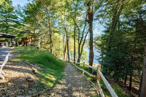 A gravel path with a wooden fence leading to a body of water, surrounded by trees and a building with a covered area. - Cozy Lakefront Retreat w/ Game Room & Hot Tub (McHenry)