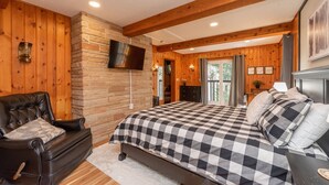 A bedroom with a checkered bedspread, a leather chair, wooden walls, and a mounted TV. - Cozy Lakefront Retreat w/ Game Room & Hot Tub (McHenry)