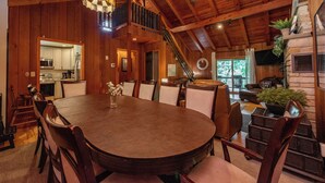 A wooden interior with a large round dining table, chairs, a staircase, and a kitchen visible through an open doorway. - Cozy Lakefront Retreat w/ Game Room & Hot Tub (McHenry)