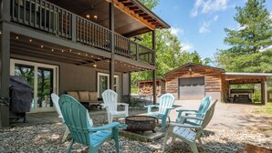A cabin with a covered deck, outdoor seating with Adirondack chairs, a fire pit, and a garage. - Cozy Lakefront Retreat w/ Game Room & Hot Tub (McHenry)