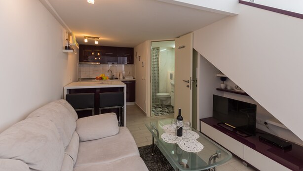 Un espace de vie compact comprenant un canapé, une table basse en verre avec une bouteille et des verres, un téléviseur et un coin cuisine avec des placards et un évier.