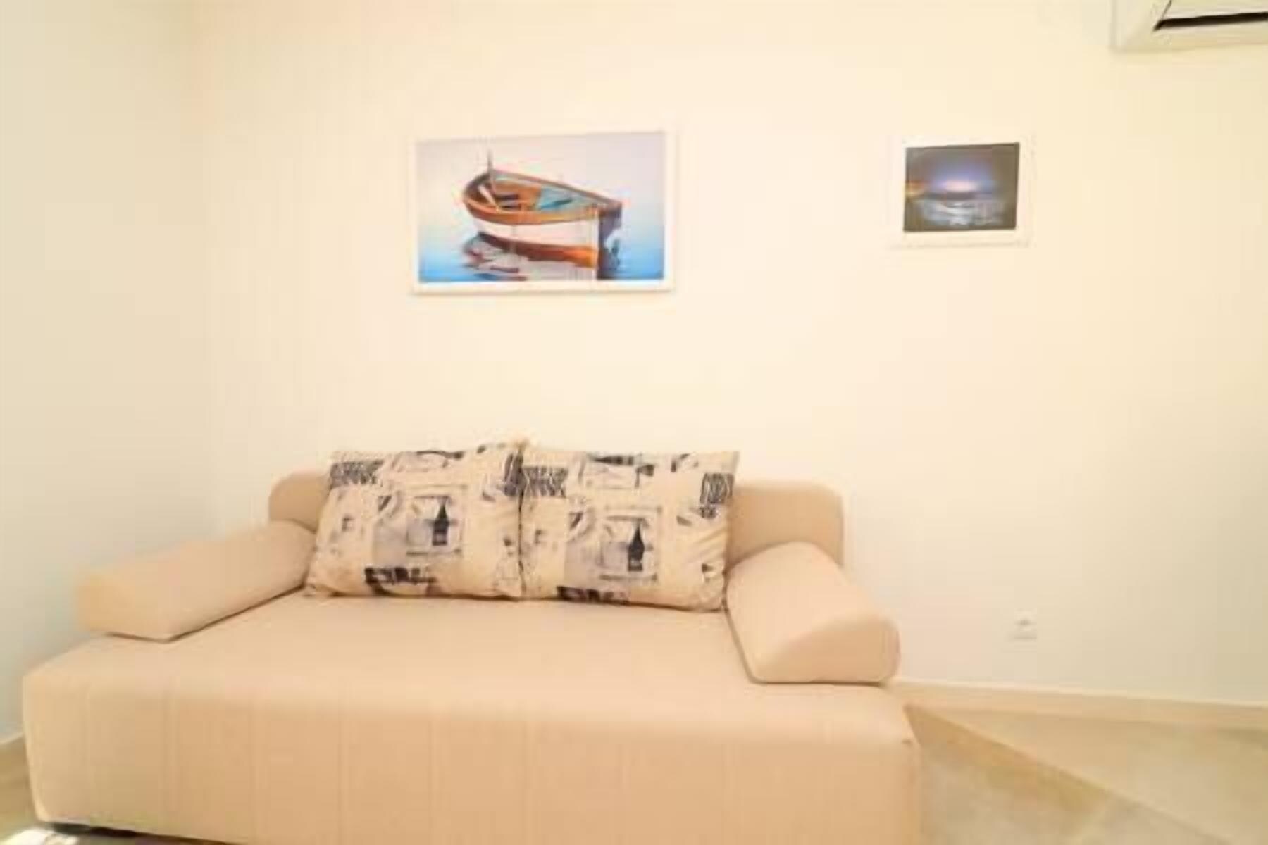 A beige sofa with patterned cushions in a room with white walls. There are two framed pictures on the wall, one of a boat and another of a sunset over water.