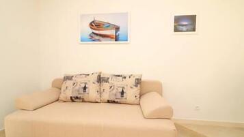 A beige sofa with patterned cushions in a room with white walls. There are two framed pictures on the wall, one of a boat and another of a sunset over water.