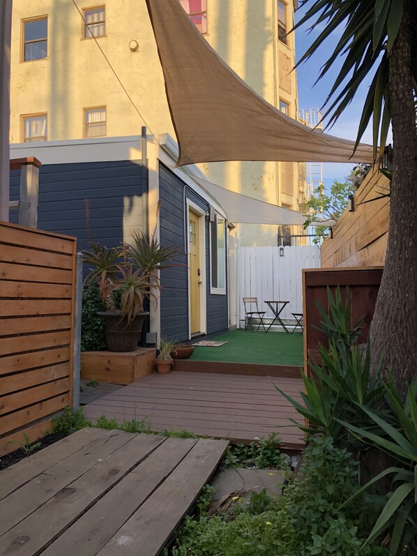A courtyard with a wooden deck, potted plants, a white fence, and a building with a dark facade and windows. - Classy Cottage (Oakland)