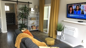 Ett vardagsrum med en grå soffa, en tv-apparat som visar ett nyhetsprogram, en bokhylla, en växt och en skjutdörr i glas.