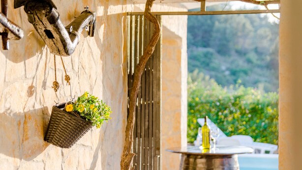Un muro in pietra con un cancello in metallo, un cesto pensile con fiori, e vista su una piscina e vegetazione.