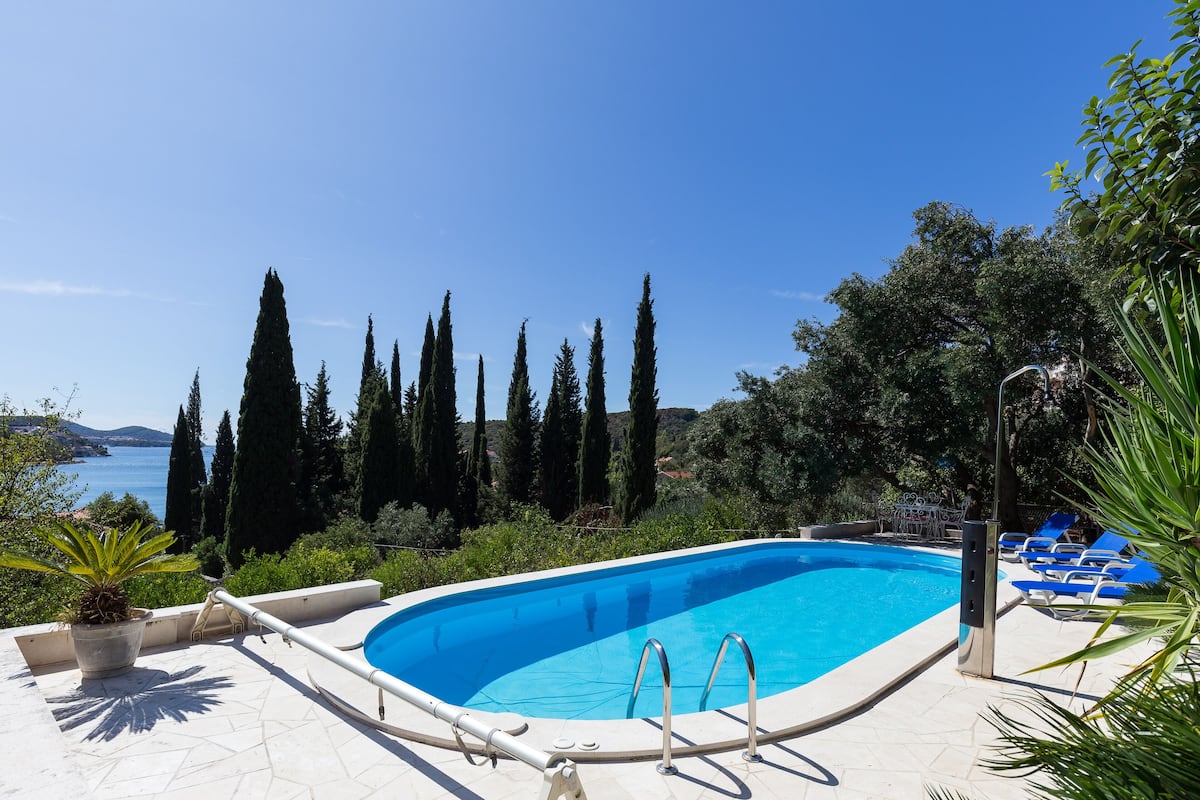 A swimming pool with clear blue water, surrounded by a white tiled deck, and overlooking a scenic view of the sea and tall trees.