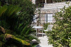 Un patio con mesa y sillas blancas, rodeado de abundante vegetación y un edificio de piedra con contraventanas blancas.